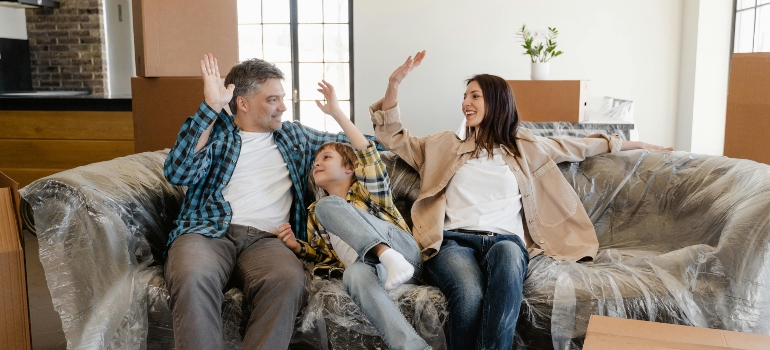 Smiling family sitting on a plastic-covered couch, surrounded by unpacked boxes in their living room.